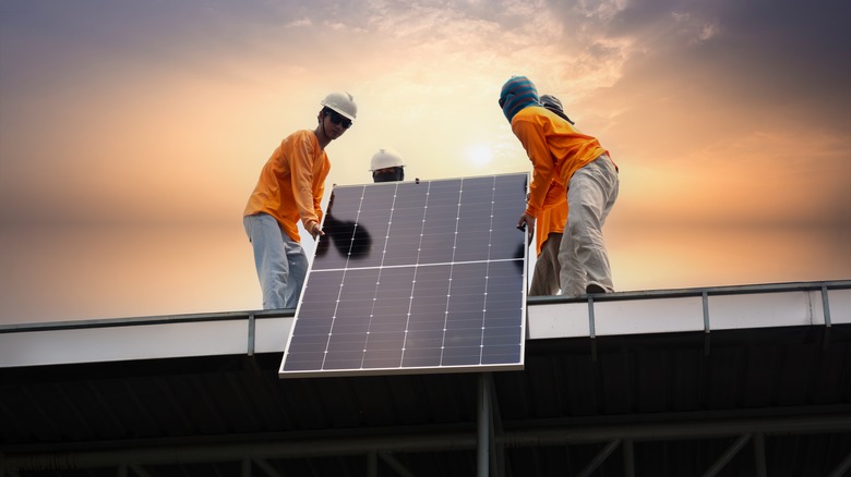 Workers in orange shirts and khaki pants install solar panels atop a building at sunset