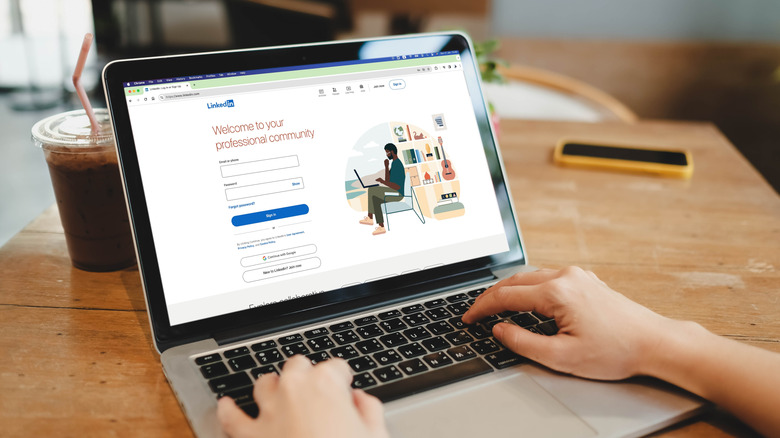 A laptop showing the LinkedIn login page is on a wooden table next to a drink in a plastic container. Hands typing on the keyboard are visible.