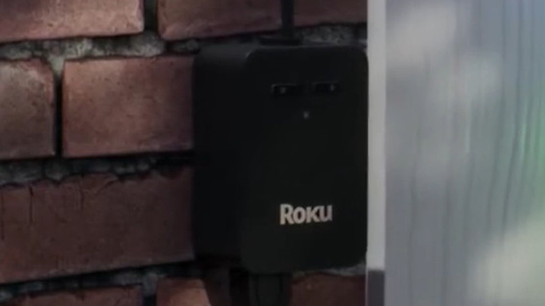 A Roku outdoor smart plug attached to a wall next to a white plank