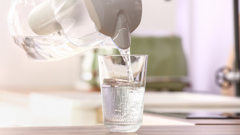 Pouring of water from modern filter jug into glass on kitchen counter, closeup